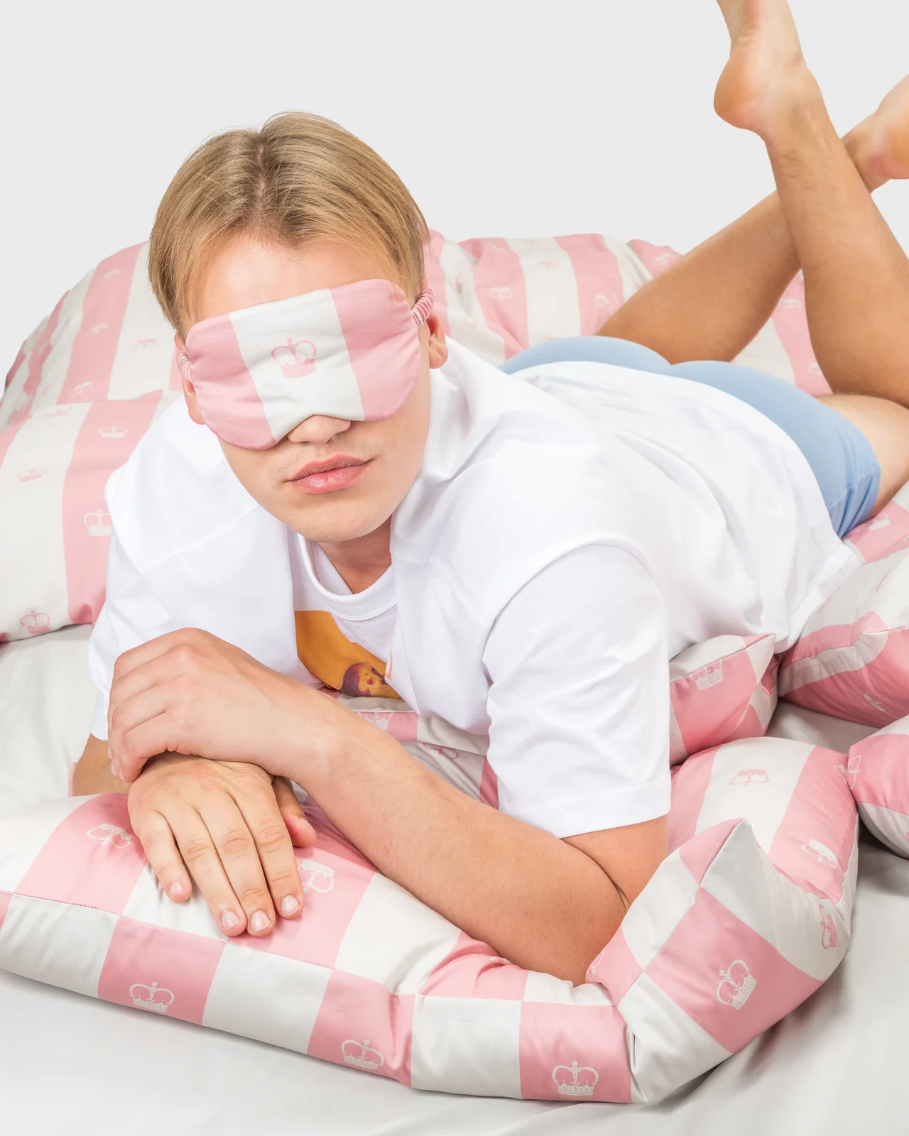 Vaikinas su rausvais miego akiniais guli ant rausvos dryžuotos patalynės „Jūsų Didenybei“