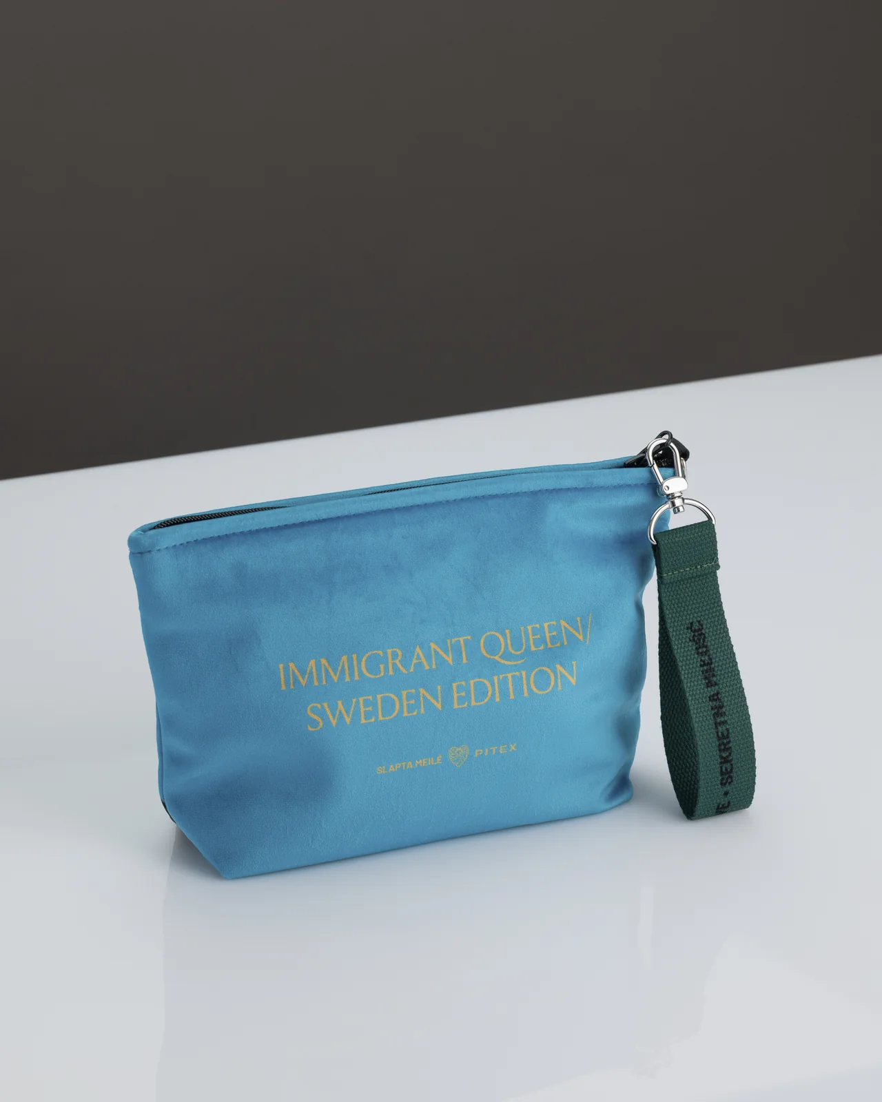 Mėlyna kosmetinė „IMMIGRANT QUEEN: Sweden Edition“ ant stalo, matoma pusė su auksiniu užrašu