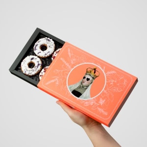 Ranka laiko atidarytą oranžinę dėžutę „Karalienės Barboros“ meduolių su glaistytais žiedais