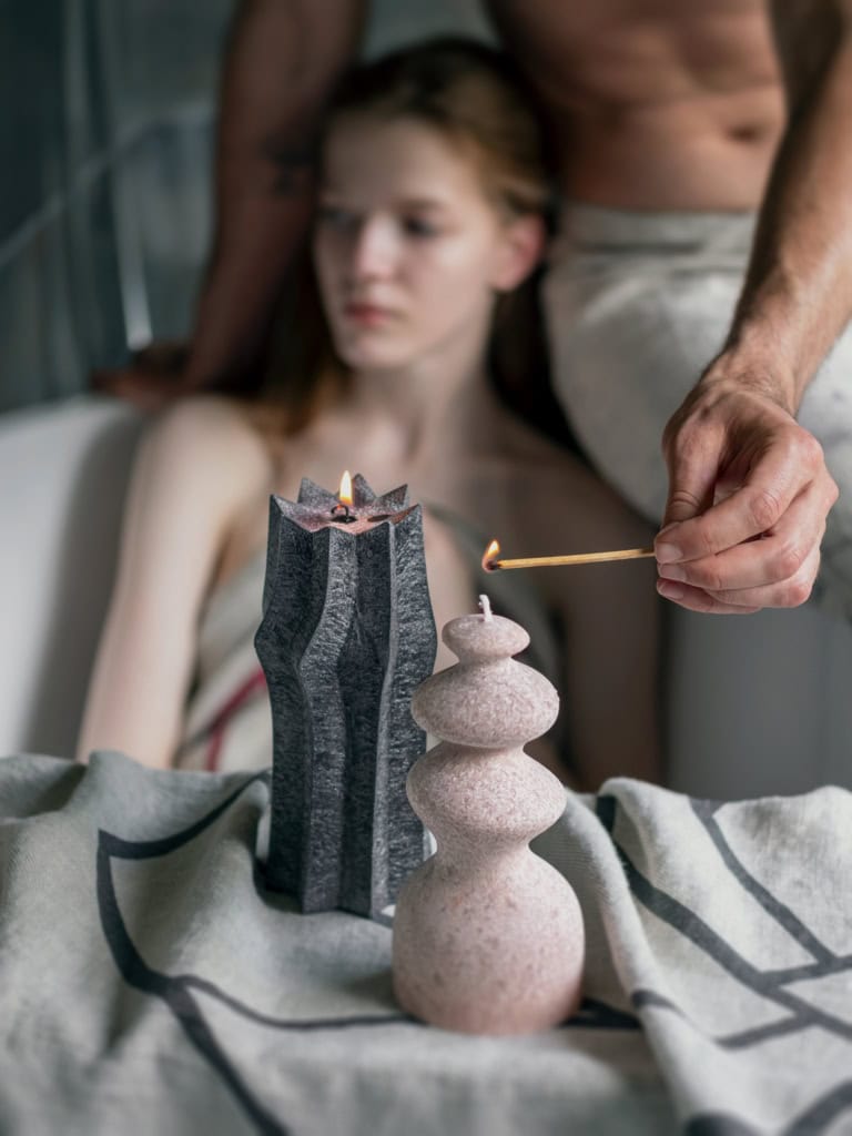 Vyras uždega pudros spalvos žvakę „Karalienė Barbora“ šalia vonioje sėdinčios moters