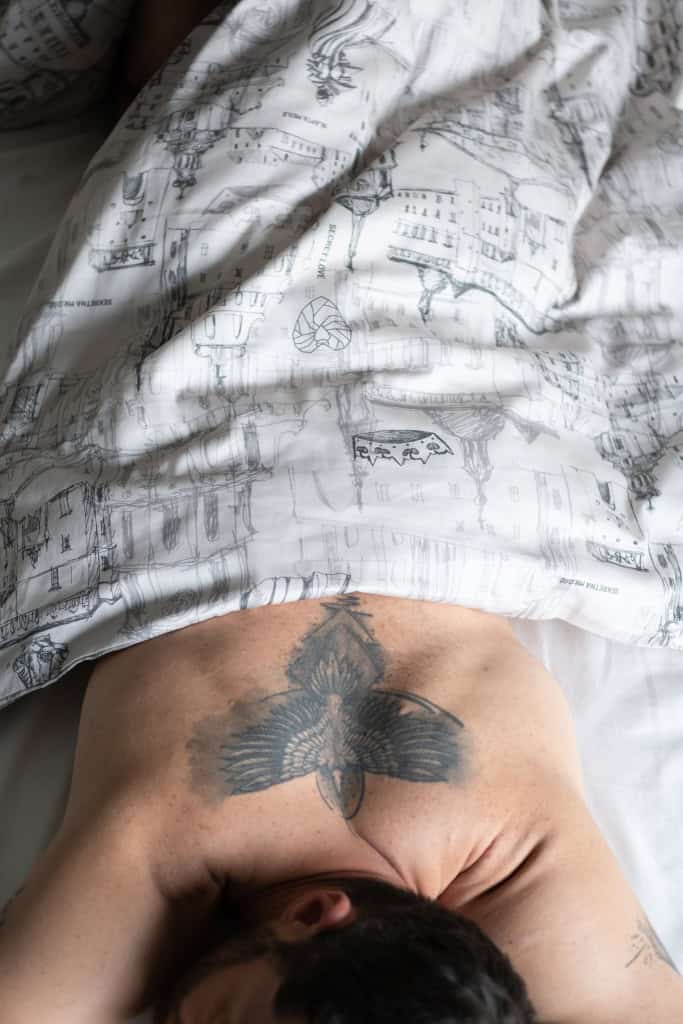 Vyras tatuiruota nugara guli po balta patalyne „Karališko Vilniaus meilės istorija“, vaizdas iš viršaus