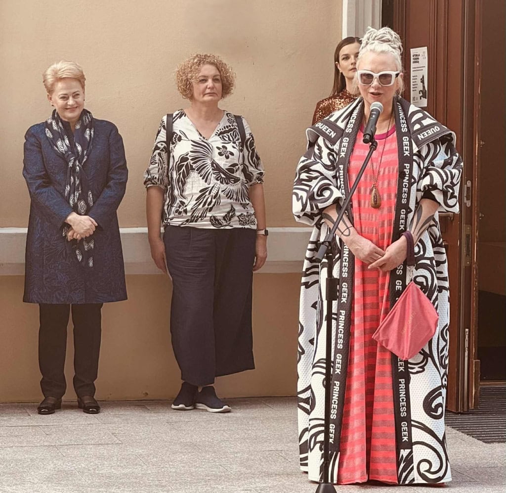 Dizainerė Julija Janus margu apsiaustu kalba, šalia stovi dvi moterys