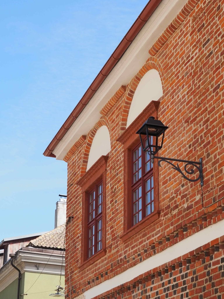 Raudonų plytų namo fasadas su arkiniais langais ir žibintu – ekskursija „Karaliaus Augusto laiškai“