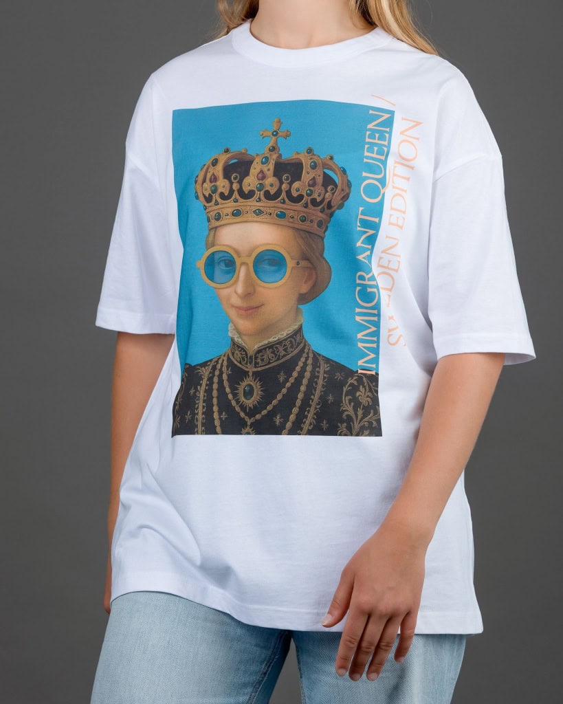 Moteris su baltais marškinėliais „IMIGRANT QUEEN / SWEDEN EDITION“ ir džinsais, ranka kišenėje