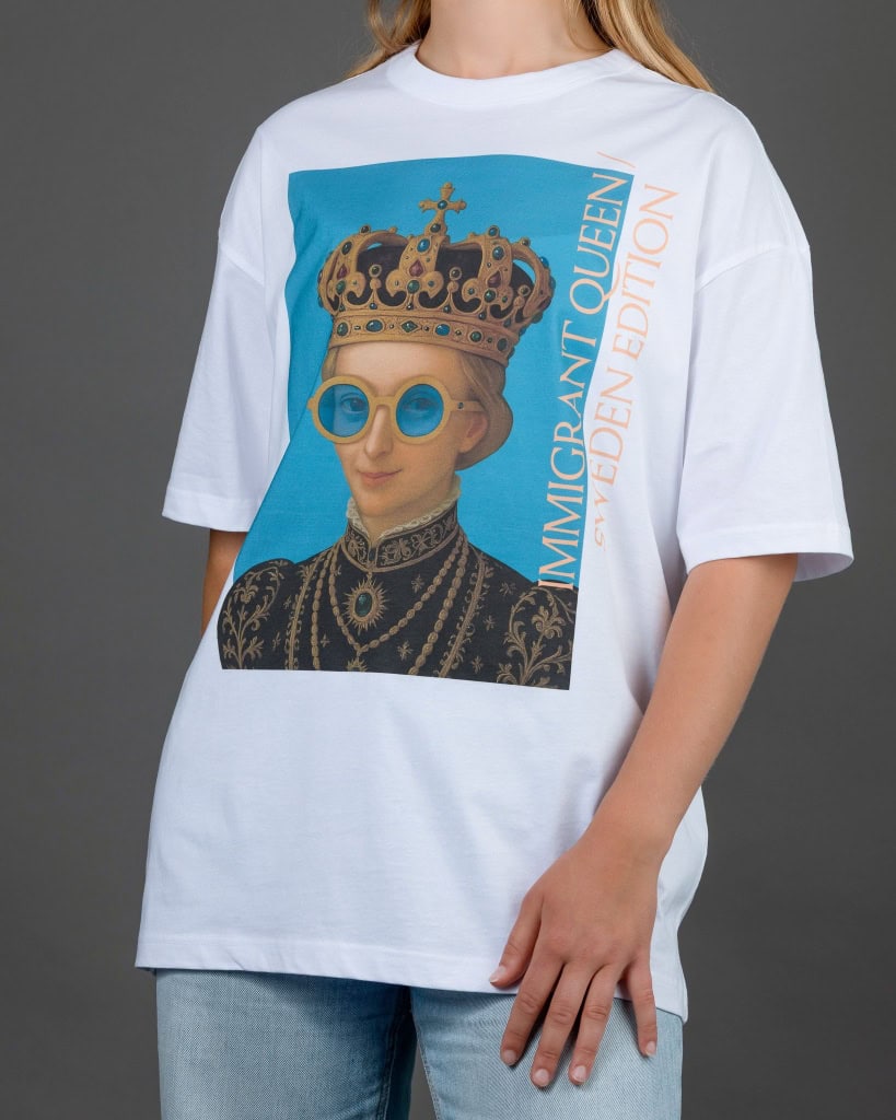 Moteris su baltais marškinėliais „IMIGRANT QUEEN / SWEDEN EDITION“, rankos nuleistos
