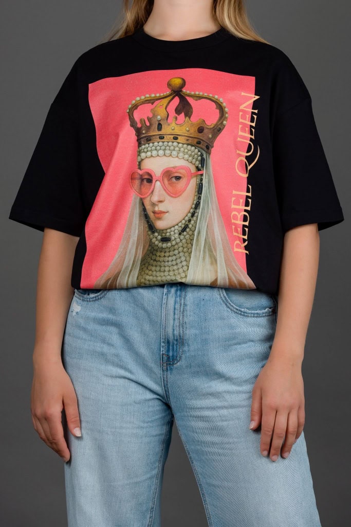 Moteris su juodais marškinėliais „REBEL QUEEN“ ir šviesiais džinsais, ranka kišenėje