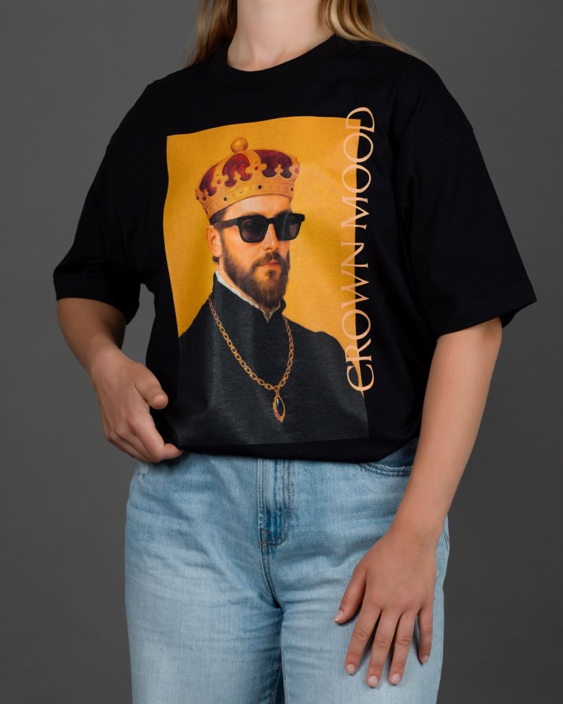Moteris su juodais marškinėliais „CROWN MOOD“ ir šviesiais džinsais, ranka kišenėje