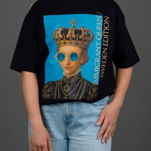 Moteris su juodais marškinėliais „IMIGRANT QUEEN / SWEDEN EDITION“ ir džinsais, ranka ant klubo