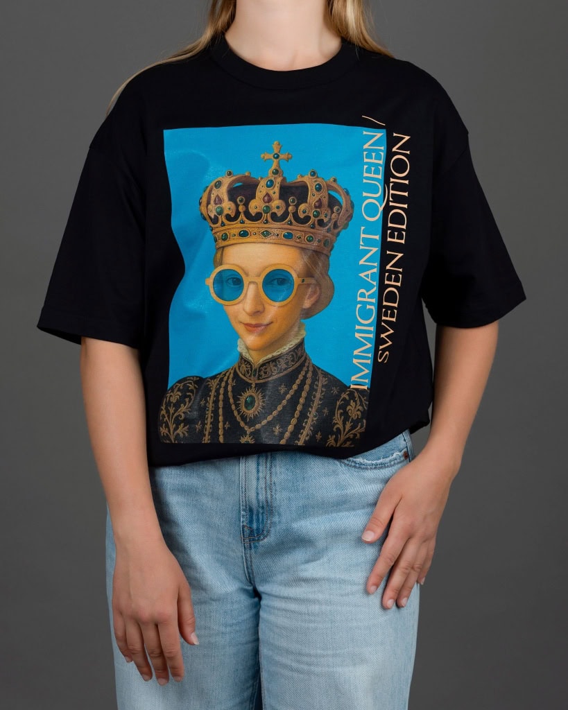 Moteris su juodais marškinėliais „IMIGRANT QUEEN / SWEDEN EDITION“ ir džinsais, ranka ant klubo