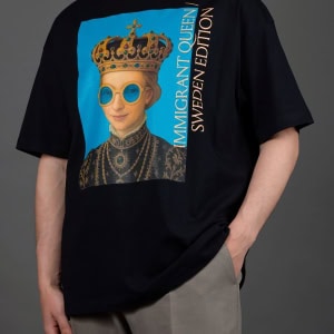 Vyras su juodais marškinėliais „IMIGRANT QUEEN / SWEDEN EDITION“, ranka kišenėje