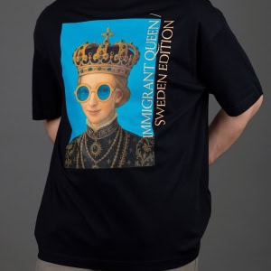 Vyras su juodais marškinėliais „IMIGRANT QUEEN / SWEDEN EDITION“, rankos už nugaros