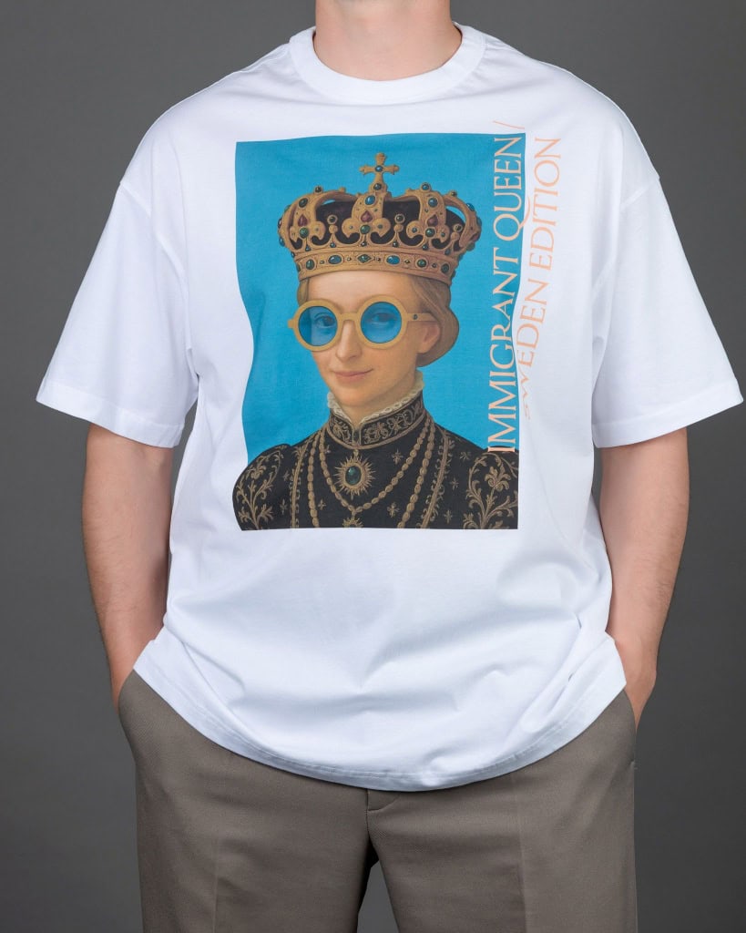 Vyras su baltais marškinėliais „IMIGRANT QUEEN / SWEDEN EDITION“, rankos kišenėse