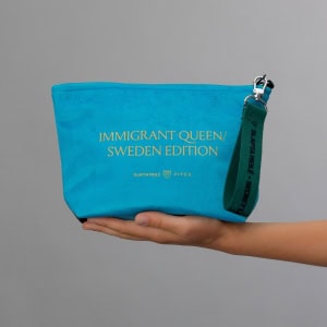 Mėlynos kosmetinės „IMMIGRANT QUEEN: Sweden Edition“ nugarėlė su auksiniu užrašu ant delno