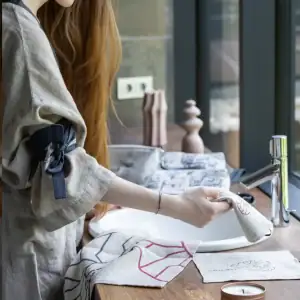 Moteris lininiu chalatu prie praustuvės su veido rankšluostėliais „Slapta meilė“