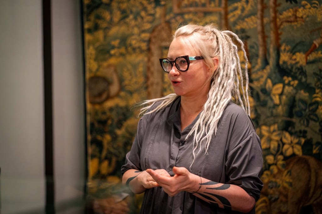 Julija Janus su šviesiomis kasytėmis kalba parodoje prie gobeleno