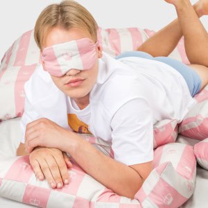 Vaikinas su rausvais miego akiniais guli ant rausvos dryžuotos patalynės „Jūsų Didenybei“