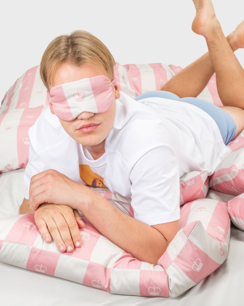 Vaikinas su rausvais miego akiniais guli ant rausvos dryžuotos patalynės „Jūsų Didenybei“