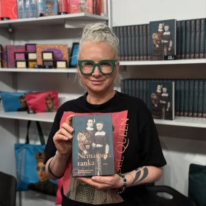 Moteris žaliais akiniais laiko knygą „Nematoma ranka“ prie knygų stendo