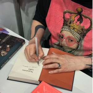 Ranka rašo dedikaciją atverstoje knygoje „Nematoma ranka“ ant stalo