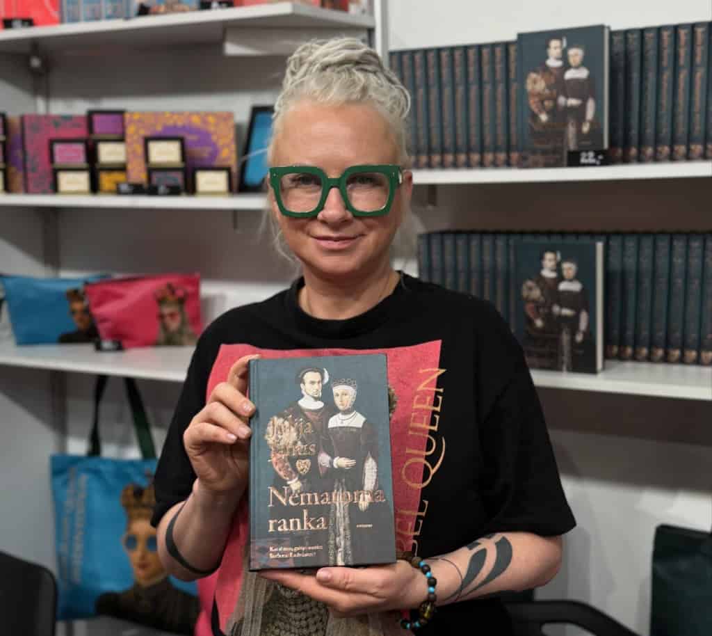 Julija Janus juodais akiniais laiko knygą „Nematoma ranka“ knygyne prie lentynų